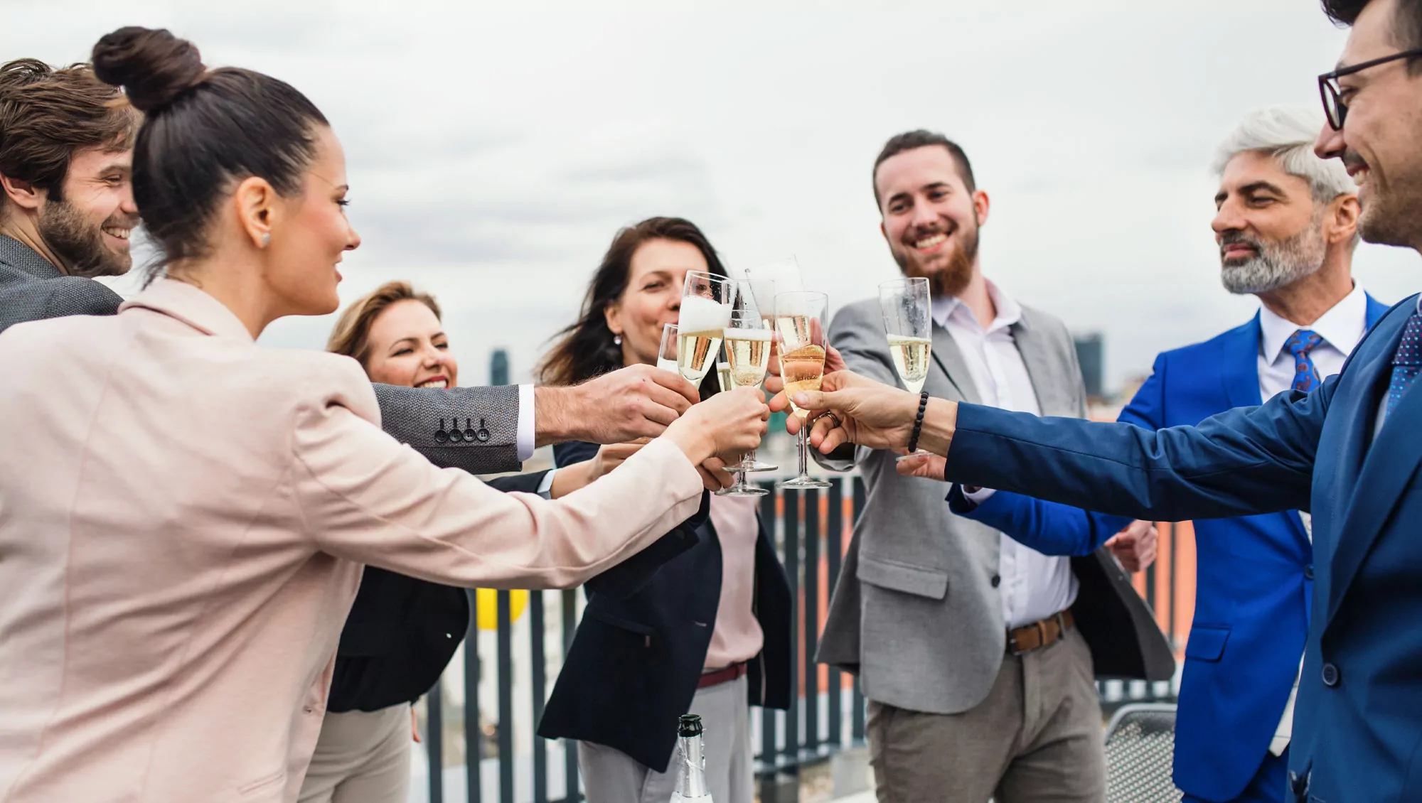 Group dressed in suits clinks champagne flutes while standing along a fence